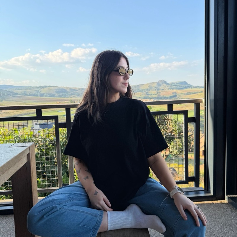 Kels in round sunglasses sitting cross-legged on a bench, looking out over green hills.