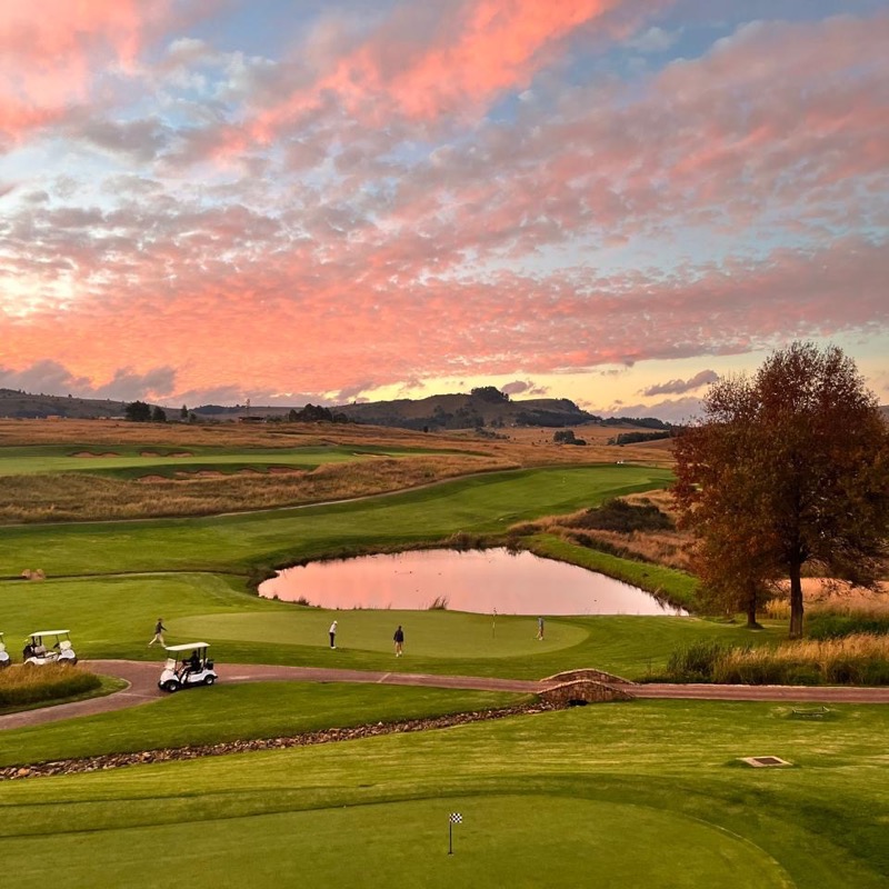 Pink sunset clouds over a golf course with a pond and rolling hills.